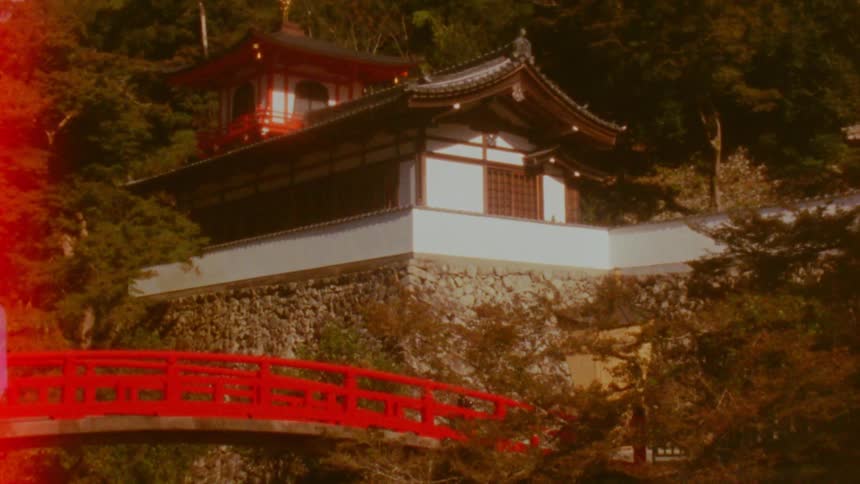 A traditional japanese building with a curved roof stands on a stone foundation with a vibrant red bridge