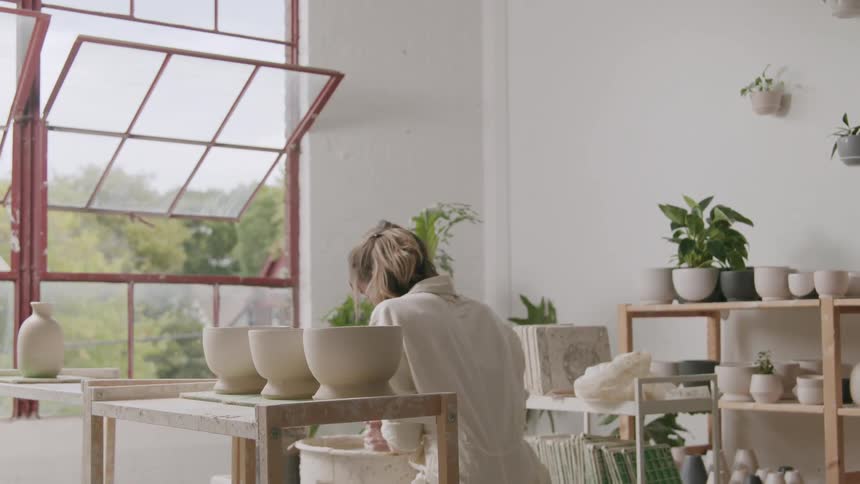 A person is working on pottery in a welllit studio with large windows surrounded by various potted plants and ceramic pieces on shelves and tables