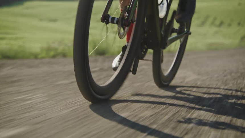 Close up on the wheels of a gravel bike being ridden at high intensity up a dirt road surrounded by green fields and trees in the background