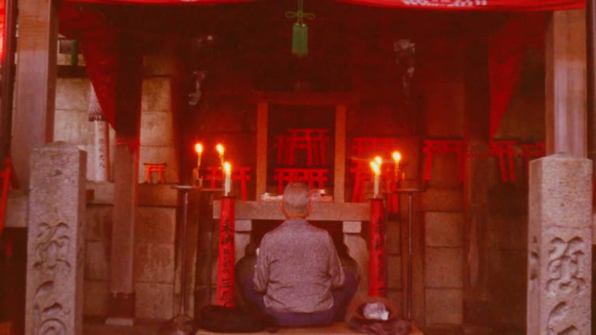 A man kneels in prayer before a small japanese shrine illuminated by flickering candles