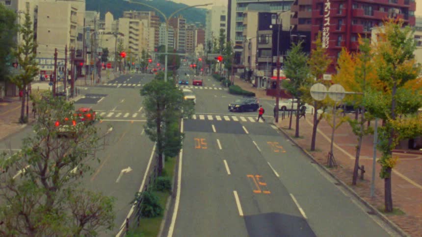 A wide multilane street cuts through a densely populated urban area with pedestrians crossing at a crosswalk