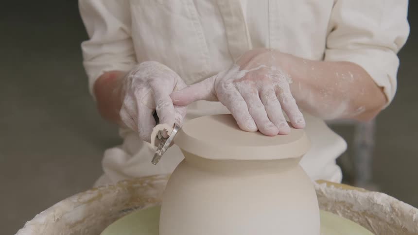 A person is shaping a clay pot on a pottery wheel using a tool to refine the top edge with hands covered in clay