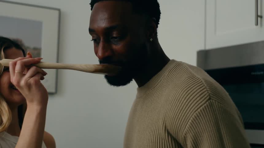 Blonde white woman feeding food from a spoon to a black man while they both cook and laugh at a kitchen indoors