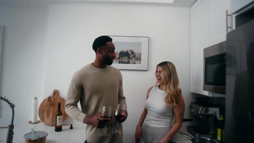 Smiling black male and white blonde female standing up at a kitchen indoors pouring glasses of wine and toasting with the glasses