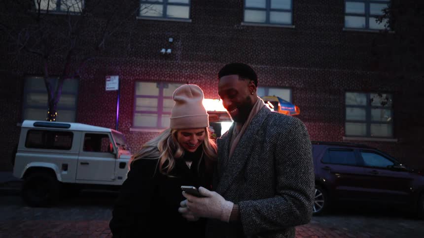 Black man and white blonde woman wearing winter clothes standing outdoors on the streets of a suburban area looking at a smart phone while they talk and laugh during night time