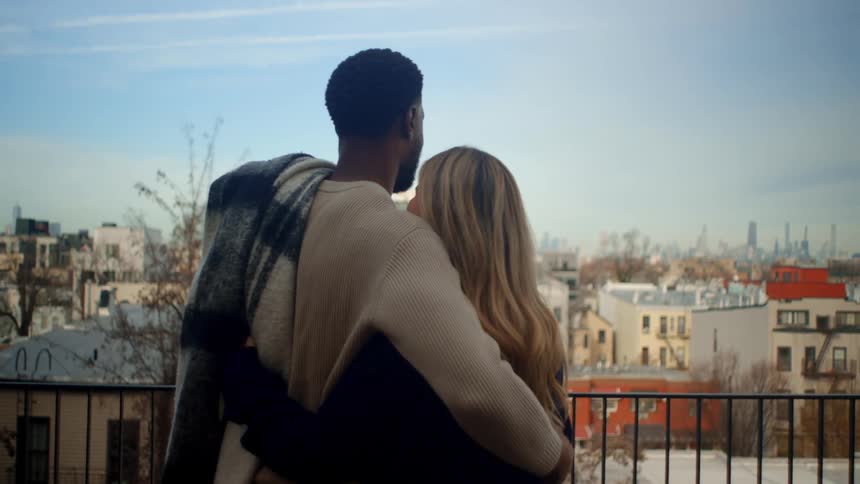 Blonde white woman and black man wearing winter clothes outdoors hugging and smiling while looking at the view of a suburban area during day time