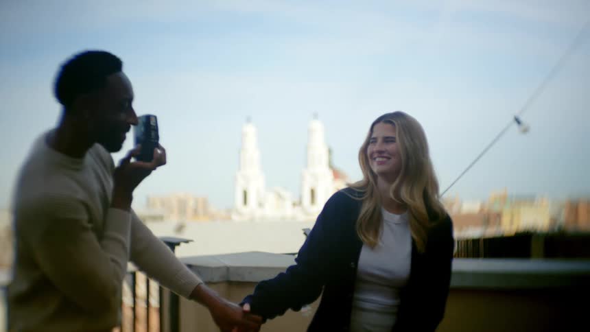 A couple holds hands as the man photographs the woman with a city backdrop