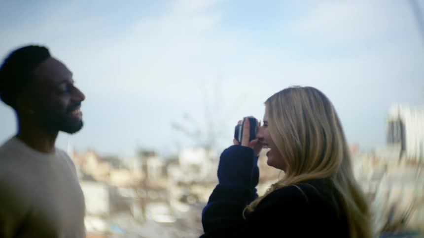 White blonde woman photographing a black man while both laugh outdoors with a suburban area on the background during day time