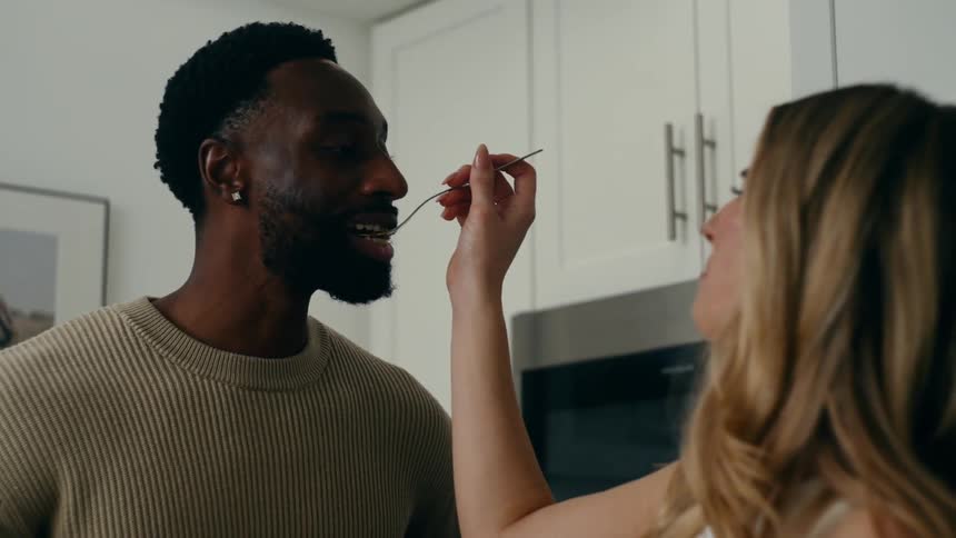 Woman offers food with a spoon to a man in a kitchen