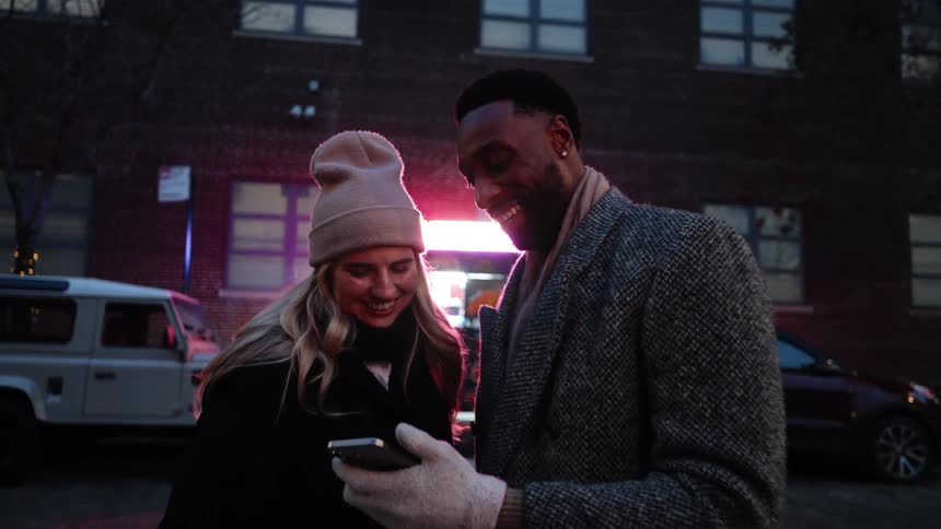 White blonde woman and black man standing outdoors looking at a smart phone and laughing during night time in the winter