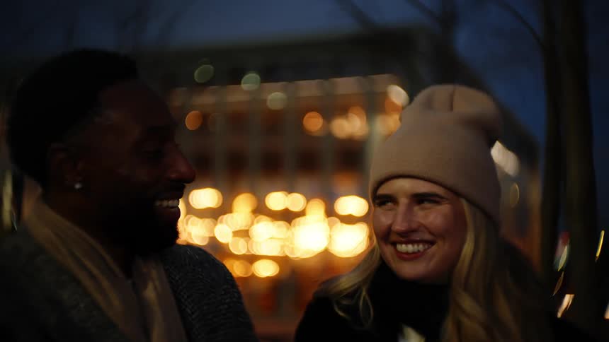 Man and woman laugh while standing outdoors near a brightly lit building at night