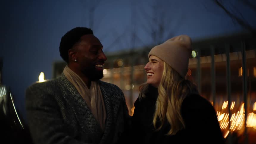 Two people stand closely together outdoors at night laughing with amusement park lights in the background