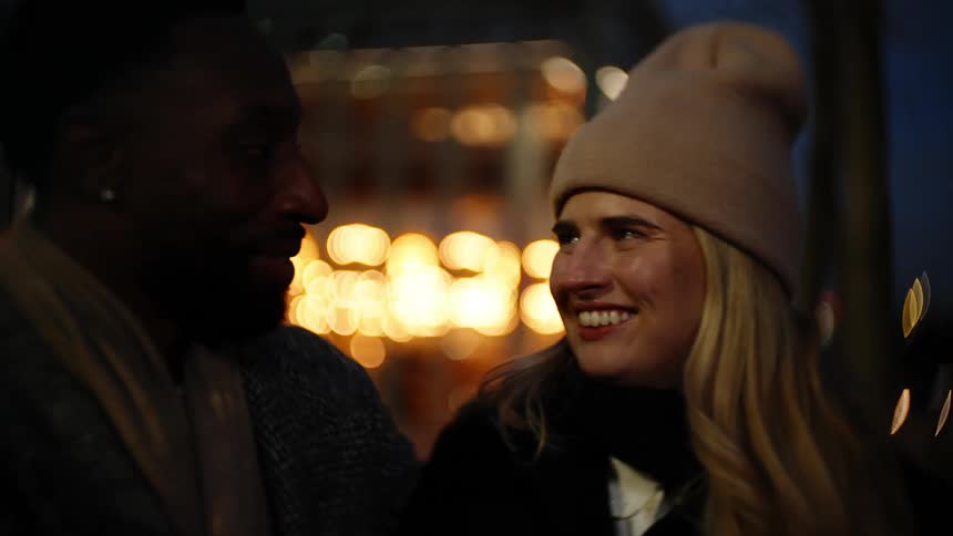 A woman wearing a beanie and is smiling while talking to a man who is next to her and there are several blurred lights passing behind them