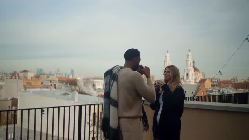A man and woman drink from mugs on the balcony