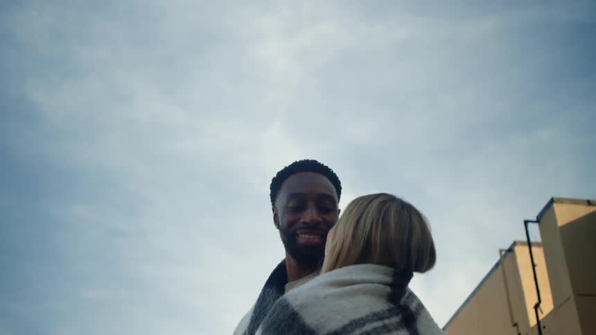 A man and a woman are wrapped in a blanket and look at each other and kiss on a clear day