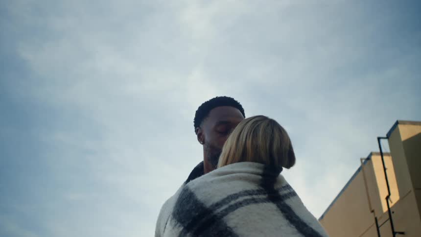 A man and woman hug and kiss with a blanket wrapped around them outside
