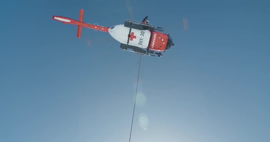 Two people next to a rope that is hanging from an orange rescue helicopter flying in the blue sky in a snowy region
