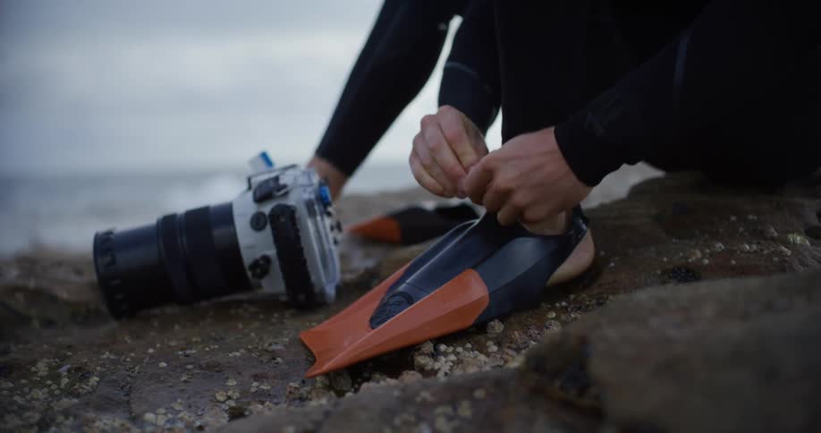 A person wearing a wetsuit fastens orange fins while sitting on a rocky beach with a camera nearby