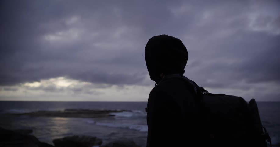 Silhouette of a person wearing a sweatshirt and carrying a backpack enjoying the view by the sea on a cloudy morning