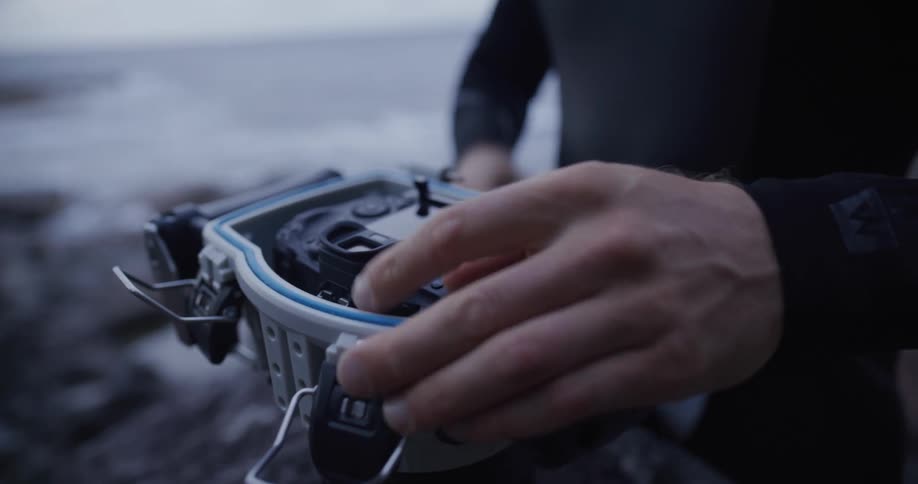 Man wearing a wet suit while standing at the coast places a camera in a waterproof case
