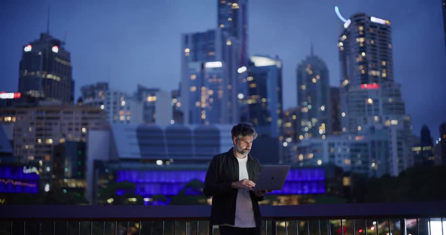 A grayhaired man is fiddling with a laptop while he is standing and behind there is a big city with tall buildings in a dusk