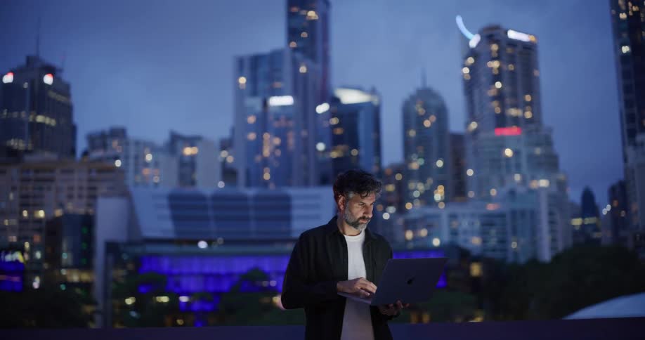 Man outdoors using a laptop computer standing up on a balcony with a metropolitan area in the background during dusk or dawn