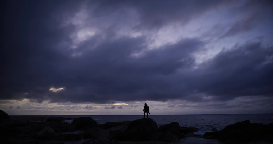 A man stands on rocks of the shore under heavy clouds
