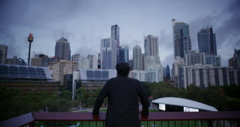 White man standing on a terrace outdoors looking at a metropolitan area surrounded by trees and nature during day time