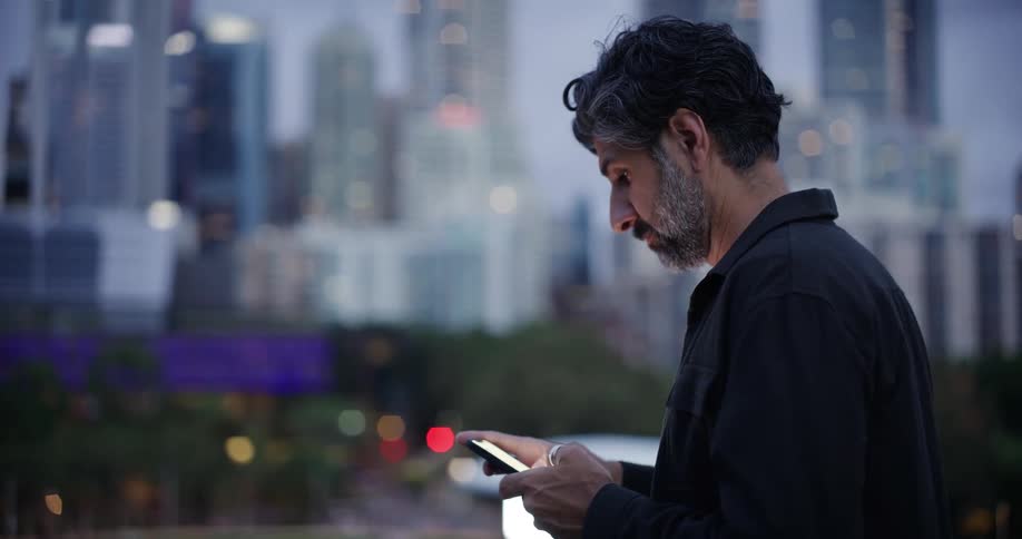 A man with dark hair and a beard looks down while using his phone with a blurred city skyline in the background