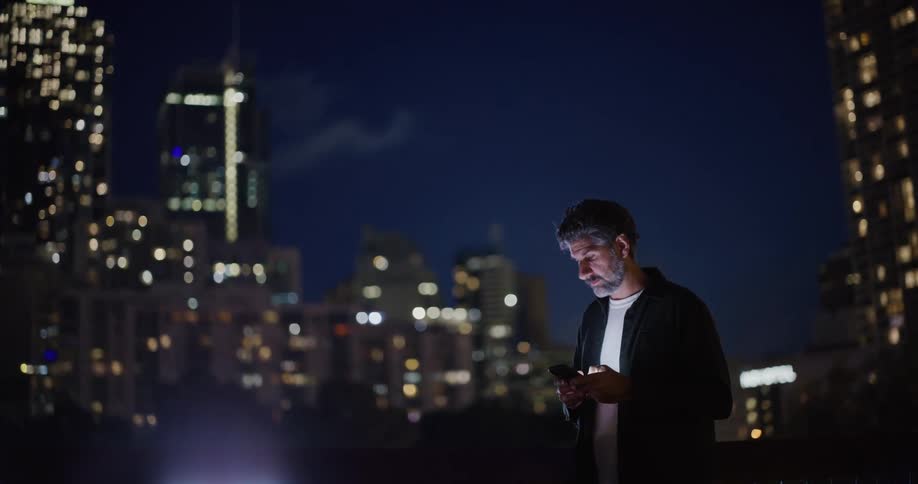 Man wearing a black shirt standing on top of a building outdoors at night time using a smartphone