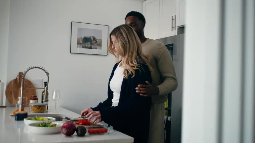 A woman cutting some vegetables on a white kitchen table and a man is hugging her from behind and they are both smiling