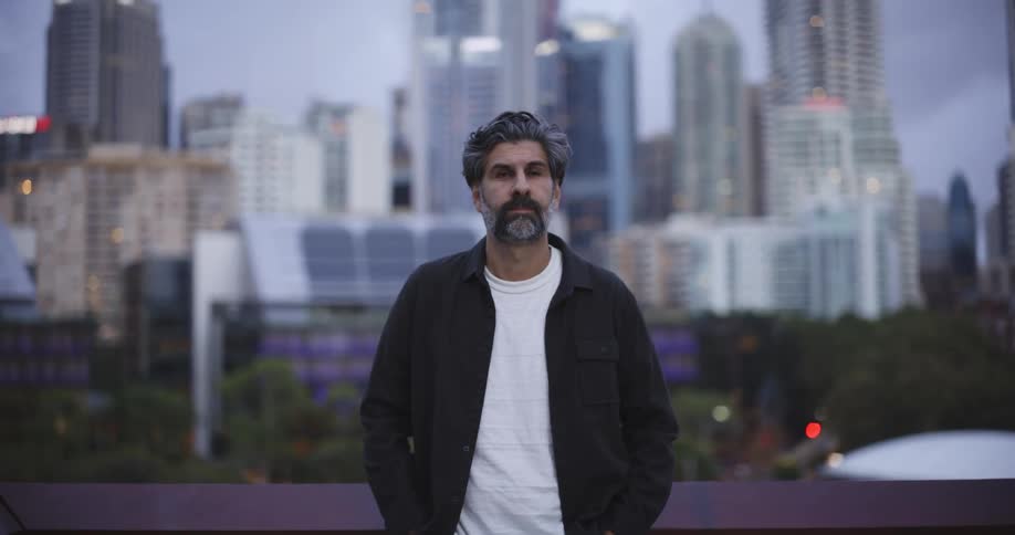 A middle aged man with graying hair and a beard stands centered against a blurred urban skyline backdrop at dusk