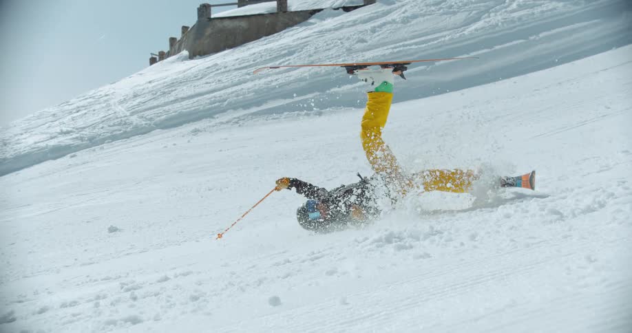 A skier in yellow pants and a black jacket is falling on a snowy slope with skis in the air and snow flying around
