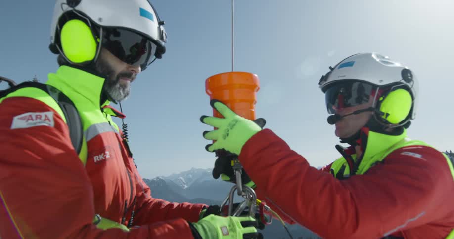 Two people in red and yellow rescue gear are preparing an orange container attached to a rope in a snowy mountain environment
