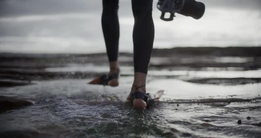 A person wearing black leggings and orange swim fins walks through shallow water holding a camera in one hand