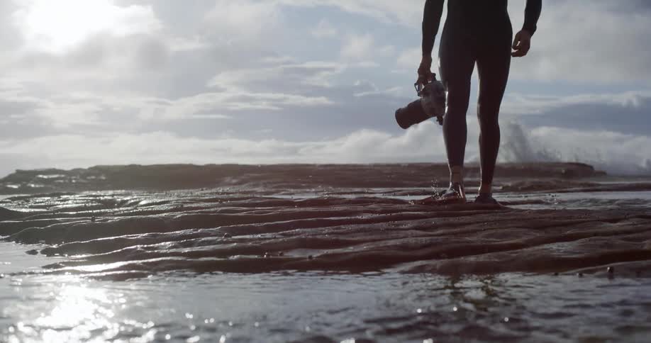 A person in a wetsuit and flippers walks on a rocky shore holding a camera with a cloudy sky