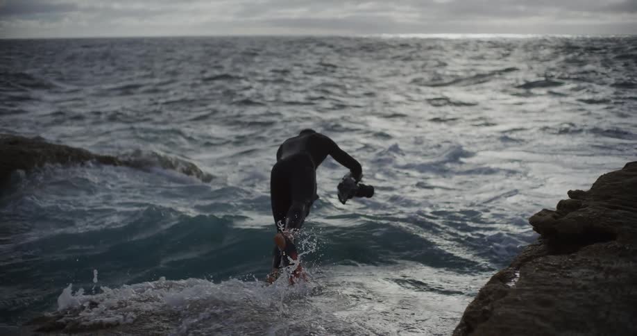 A person in a black wetsuit with flippers holding a camera enters the ocean from a rocky shoreline on an overcast day