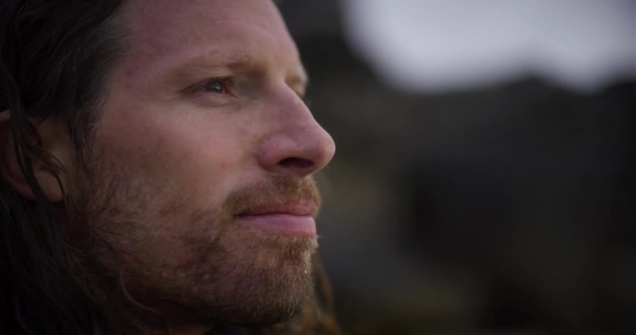 The profile of a bearded man with long hair flowing in the wind standing outdoors at dusk