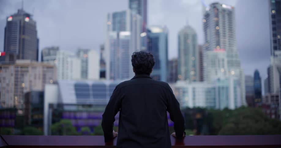 A man in a dark jacket stands at a railing looking out at a city skyline under a cloudy sky