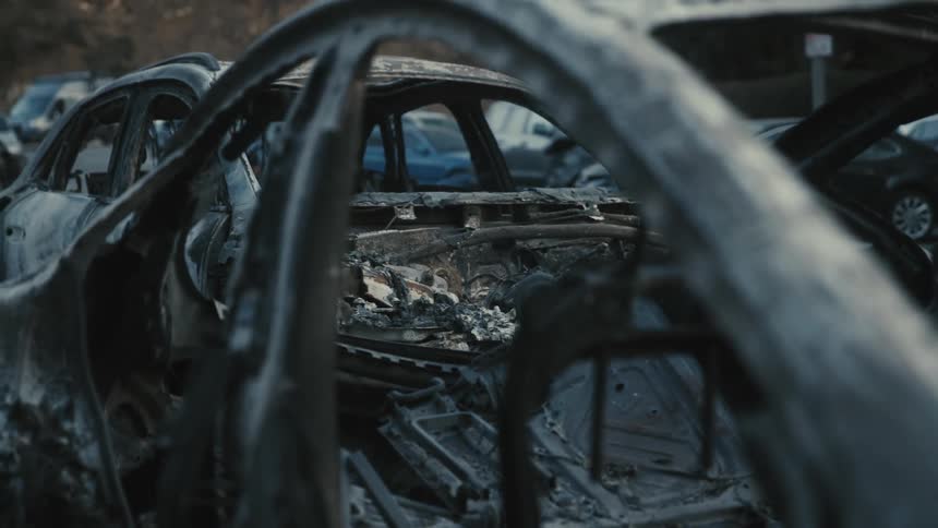 A charred and gutted vehicle is shown after being burned with other cars visible in the background