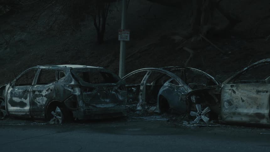 Three severely burned cars are parked on the side of a street under dim lighting