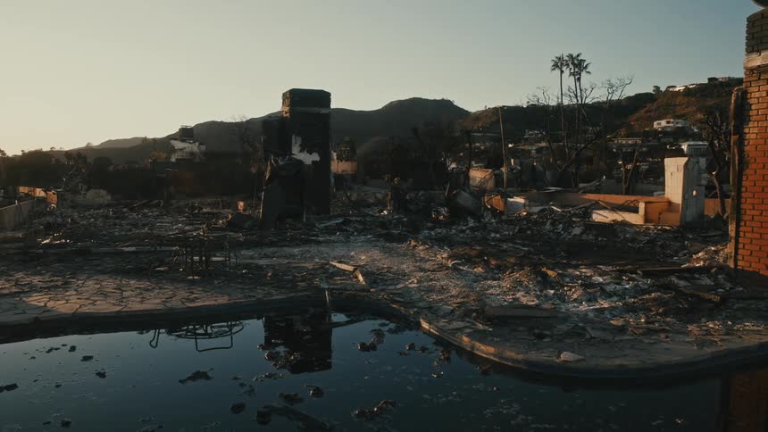 A desolate scene of a fire ravaged area with the remains of structures a water filled pool in the foreground and mountains in the background