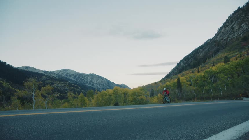 A cyclist in a red jacket rides along a road with mountainous scenery and the shot briefly shows a shadow of the bikes wheels
