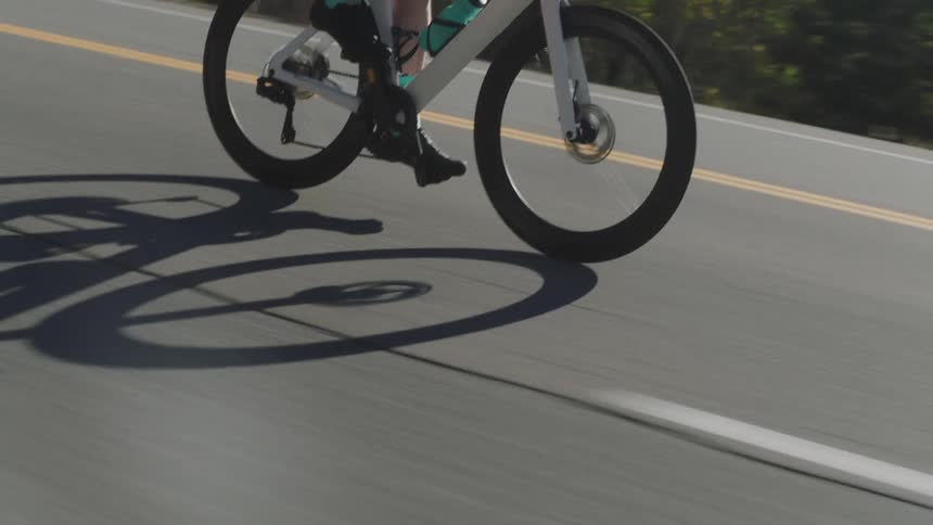 A cyclist in a red jacket rides along a road with mountainous scenery and the shot briefly shows a shadow of the bikes wheels