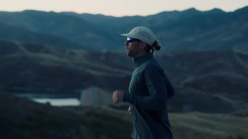 A man wearing a cap and reflective sunglasses runs against a backdrop of mountains and a body of water at dusk