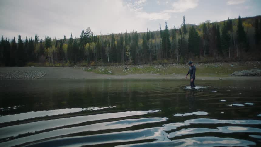 Man wearing a wet suit wades in the water near a shore covered by conifers
