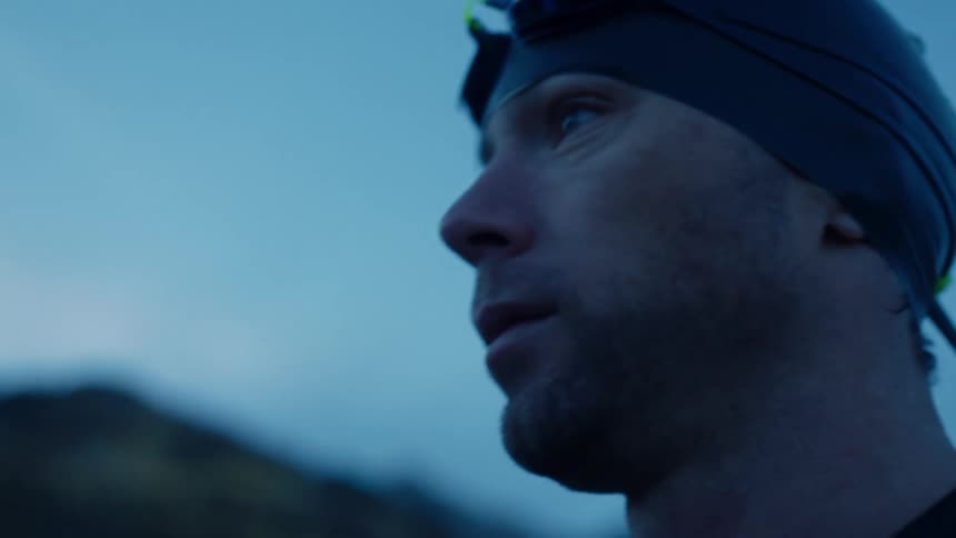 A man in a swim cap and goggles looking off to the side before adjusting his goggles in front of a blurry mountain background at dusk