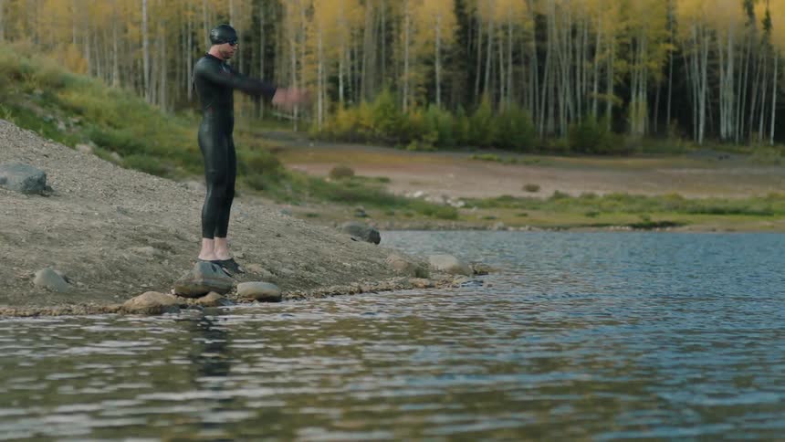 A person in a black wetsuit and swim cap stands on rocks at the edge of a lake preparing to enter the water with a backdrop of trees and a forested area