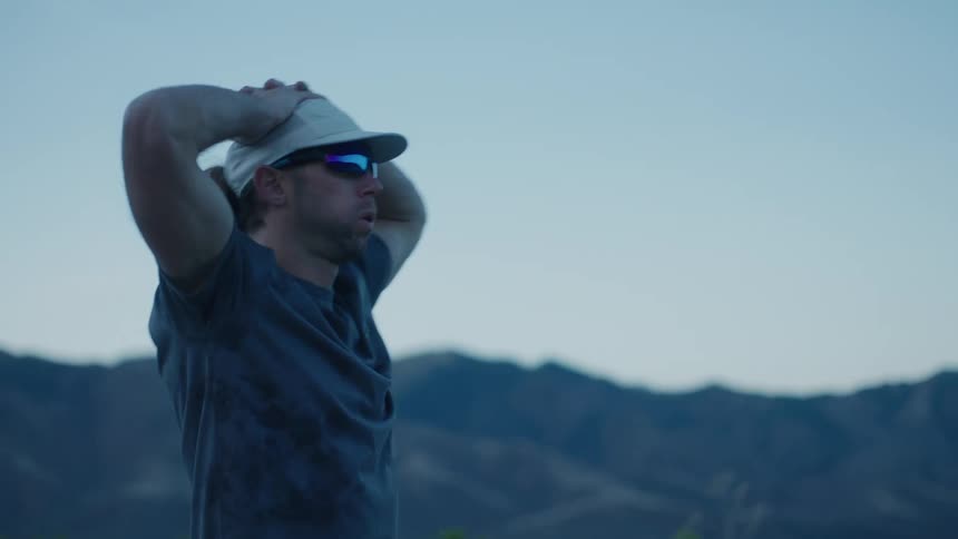 Man wearing cap and glasses with hands on head while standing in natural area with view of mountains