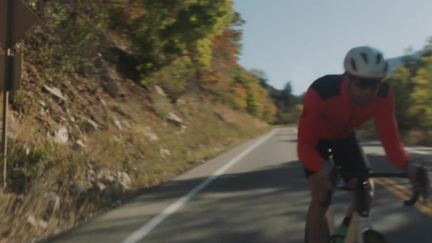 A cyclist wearing an orange and black long sleeve shirt black shorts a white helmet and red sunglasses rides a white bicycle along a paved road surrounded by trees and mountains under a bright sky
